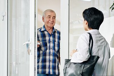 Ein älterer Mann mit grauen Haaren und einem karierten Hemd steht lächelnd an der geöffneten Tür, während er mit einer Person spricht, die eine Tasche trägt und ihm gegenübersteht.